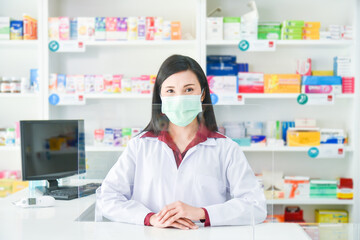 Asian professional young pharmacist wearing face mask while standing behind partition at drugstore.