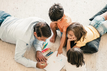 Concentrated dad and kids lying on carpet and painting doodles. Middle-aged father drawing with colorful pens and playing with cute children at home. Childhood, game activity and fatherhood concept