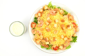 Top view of a plate of shrimp salad sprinkled with cheese crumbs and white bread croutons. A jar of white sauce is nearby