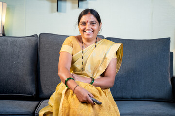 Indian woman in saree watching TV at home while sitting on sofa by holding remote during leisure...