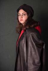 A girl in a black hat , wearing a red hoodie and a black leather jacket on the background of a dark wall.