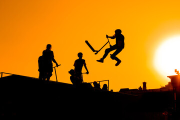 Unrecognizable teenage boy silhouette showing high jump tricks on scooter against orange sunsetwarm sky at skatepark. Sport, extreme, freestyle, outdoor activity concept