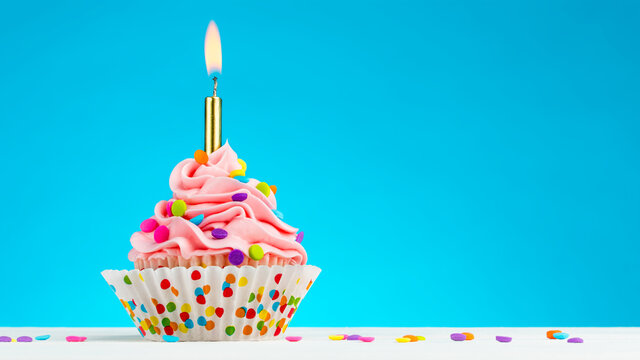Happy Birthday cupcake. Tasty cupcakes with pink cream icing and colored sprinkles. Burning candle in a cupcake. Sweet delicious dessert on white wooden table and blue background with copy space.
