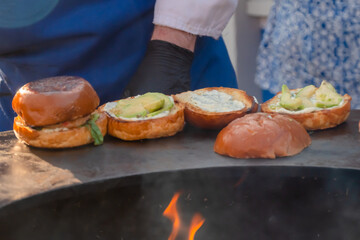 Process of preparing burgers with seafood and avocado on brazier with hot flame at summer local food market - close up view. Outdoor cooking, gastronomy, cookery, street food concept