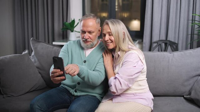 Elderly couple view photos on a smartphone sitting on the sofa in living room, positive emotions, grandfather and grandmother using smartphone.