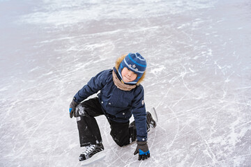 Fototapeta na wymiar cute caucasian preschooler boy learn to skate on ice rink. Kid wearing winter jacket, hood hat and neckwarmer standing on one knee. Image with selective focus