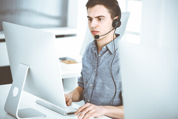 Casual dressed young man using headset and computer while talking with customers online. Call...