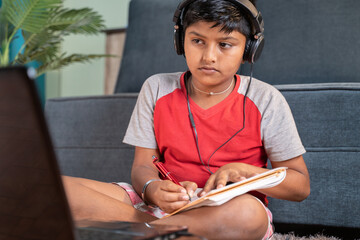 Serious kid with headphone noting down to book by seeing laptop during online class at home - concept of online classroom, online education, technology and lifestyle.