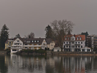 Residential buildings on the shore of Lake Constance in the center of Lindau, Germany with water reflections and bare trees in winter season on foggy day.