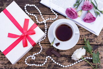 Morning time, beautiful picture for blog post in Instagram. Relax with cup of coffee with book and chocolate, decorated by flowers and beads, white gift box, enjoy your free minutes. Wooden background