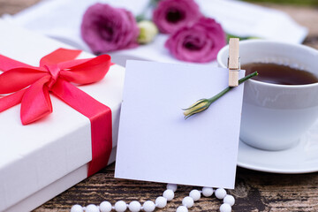 Morning time, beautiful picture for blog post in Instagram. Card for congratulations in center on clothespin, gift in white box, cup of tea, flowers on open book, beads on wooden table. Close-up