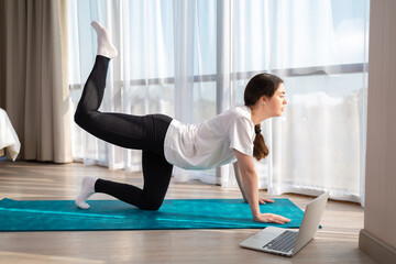 Young beautiful woman in sports clothes, doing yoga online lessons on a laptop. Indoor. Side view. The concept of online training at home