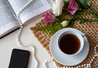 Unusual, beautiful composition for blog post  on Instagram. Cup of coffee, open book and telephone decorated with beads on white table with flowers and brown napkin background. Minute of rest.