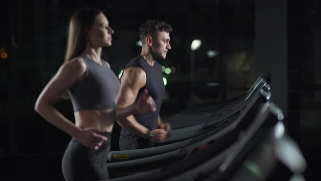 Wellness, female and man athlete run on a treadmill, two runners perform aerobic exercise and endurance training in the gym, nightlife.