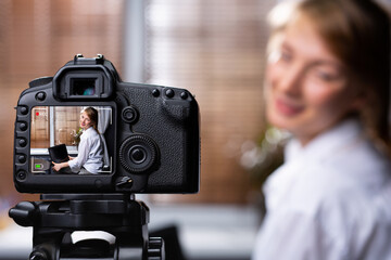 Young female blogger looking at camera and talking a video recording holding laptop at home. Social media blogger or content maker. Blonde smiling speaking in front of camera for vlog. Video tutorial