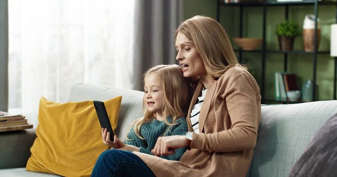 Happy mother with little daughter sitting at home on couch and talking to relatives on video call using smartphone.