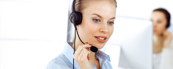 Blond woman call operator is using computer and headset for consulting clients online. Group of...