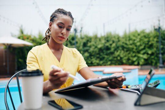 Pensive serious young Hispanic female entrepreneur sitting at table by swimming pool, reading document and taking notes - Powered by Adobe