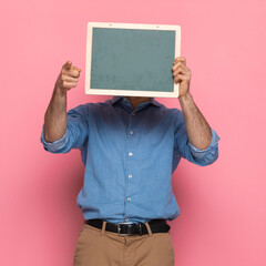 casual guy in denim shirt hiding behind blackboard and pointing finger