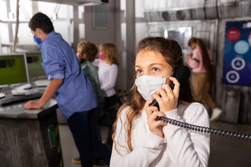 Tween girl in protective face mask talking on telephone while her friends trying to solve...