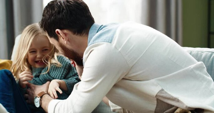 Caring father plays with little daughter sitting at home on couch and tickles her. Cute child is having fun and laughing out loud.