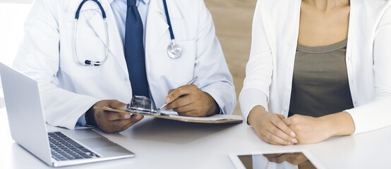 Doctor and patient discussing medical exam results while sitting at the desk in clinic, close-up. Male physician using clipboard for filling up medication history record of young woman