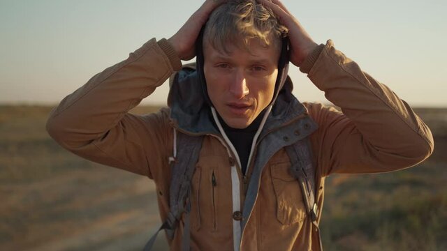 portrait of adult man traveler looking at camera at sea beach on sunrise. guy in casual jacket takes off hoodie from head with wind blowing golden hair from sunlight is enjoying scenery landscape