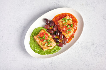 White fish with olives, pesto and sauce of tomatoes and baked peppers on white oval plate, food style concept, top view, copy space