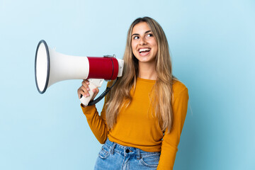 Young hispanic woman over isolated blue background holding a megaphone and looking up while smiling