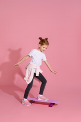 Fototapeta na wymiar cute little child girl in casual clothes riding skateboard against pink background.