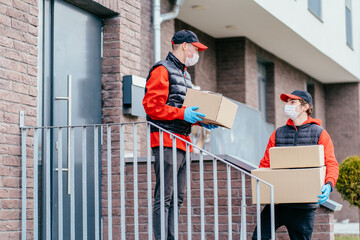 Two delivery man in masks and gloves workers in cap, red uniform walking and looking for address. Adult couriers delivering order in cardboard boxes. Delivery service, post and shipping concept