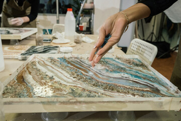 at the workshop on creating fluid art, a girl with a long manicure pours hard blue crumbs onto an epoxy painting
