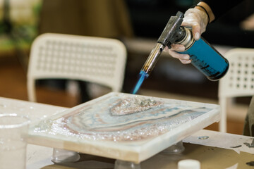 the hands of a girl in gloves working at a workshop on creating a resin painting holds a working burner in her hands