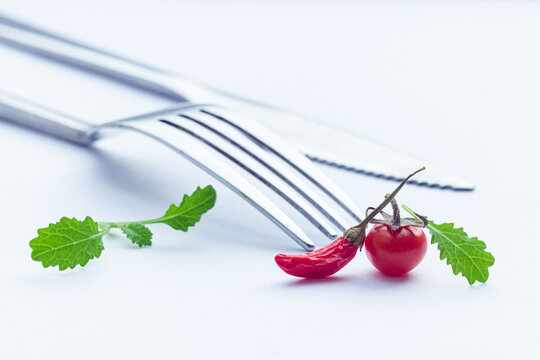 Very small cherry tomato, red chili pepper and mustard leaves on white table with fork and knife on the background. Homegrown potted mini vegetables. Tomberry tomato and tiny pepper