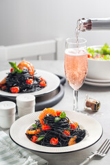 Italian dinner. Squid ink spaghetti, with shrimp and cherry tomatoes and flowing rose wine. On white table.