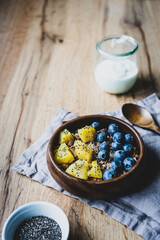 Müsli mit Heidelbeeren, Ananas und Chiasamen in einer Holz Schüssel. Joghurt im Glas, rustikaler Tisch, gesundes Frühstück.