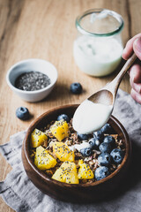 Hand gibt Joghurt über ein Müsli mit Heidelbeeren und Ananas in einer Holz Schüssel. Gesundes...