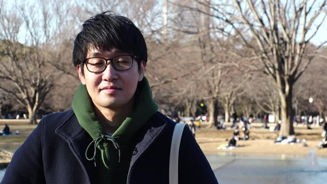 Asian (Japanese) young man sitting on the bench at the park in winter daytime. Smiling (shy smile), relaxing and looking at the camera. One person, late twenties, black hair with glasses.