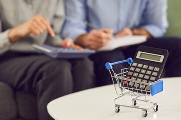 Calculator and shopping cart on table up close, people counting money in background. Cost of...