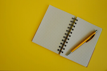 a gold colored pen rests on an open rectangular notebook on a metal spiral on a yellow background top view