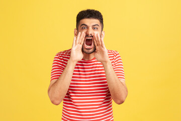 Angry nervous bearded man in striped t-shirt loudly yelling widely opening mouth holding hands on...