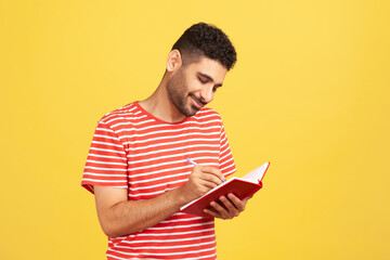 Smiling happy businessman with beard in striped t-shirt writing down to do list, making notes with pen in notebook, checking schedule. Indoor studio shot isolated on yellow background