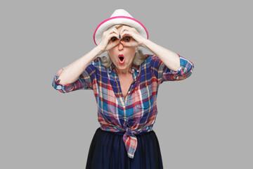 Portrait of funny surprised modern stylish mature woman in casual style with hat standing with binoculars gesture and looking with amazed face. indoor studio shot isolated on gray background.