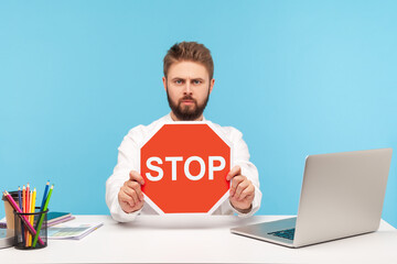 Serious bearded man office worker holding red stop sign board sitting at workplace with laptop,...