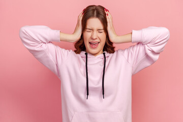 Portrait of irritated young female in hoodie covering ears and shouting with crazy expression, ignoring annoying information. Indoor studio shot isolated on pink background