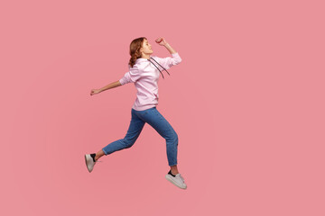 Fototapeta na wymiar Full length side view of young female in hoodie and jeans running in air, hurrying for discounts, empty copy space for advertising. Indoor studio shot isolated on pink background