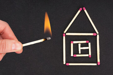 Female hand holding a lighted matchstick close to a flat house made of unused matches. Creative flat lay composition. Fire safety precautions and insurance concept.