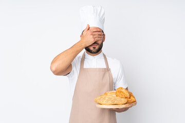 Male baker holding a table with several breads isolated on white background covering eyes by hands