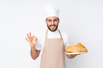 Male baker holding a table with several breads isolated on white background showing an ok sign with fingers