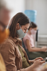 Vertical side view portrait of young woman wearing mask and using smartphone while waiting in line...